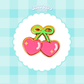 A cute enamel pin shaped like two pink heart-shaped cherries with a green bow on a scalloped white background. 