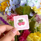 Hand holding a pink backing card with a mini heart-shaped cherry enamel pin in front of colourful flowers