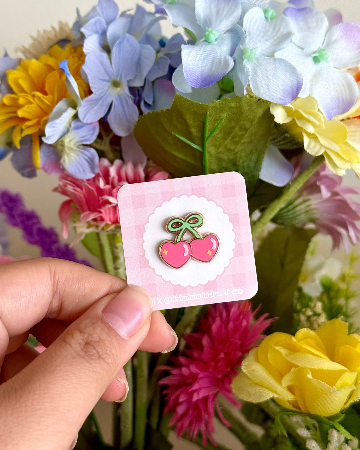 Hand holding a pink backing card with a mini heart-shaped cherry enamel pin in front of colourful flowers