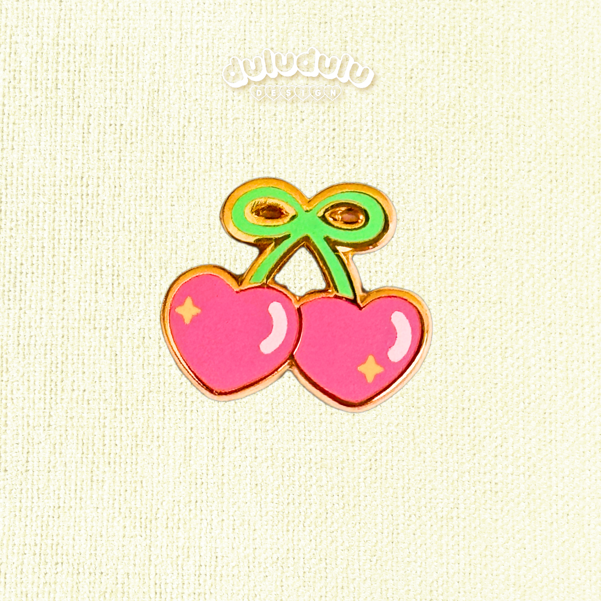 A cherry-shaped enamel pin with two pink hearts outlined in gold, green stems, and small yellow star accents, displayed on a cream-colored textured fabric background.