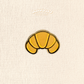 A small croissant enamel pin centered on a cream textured fabric background