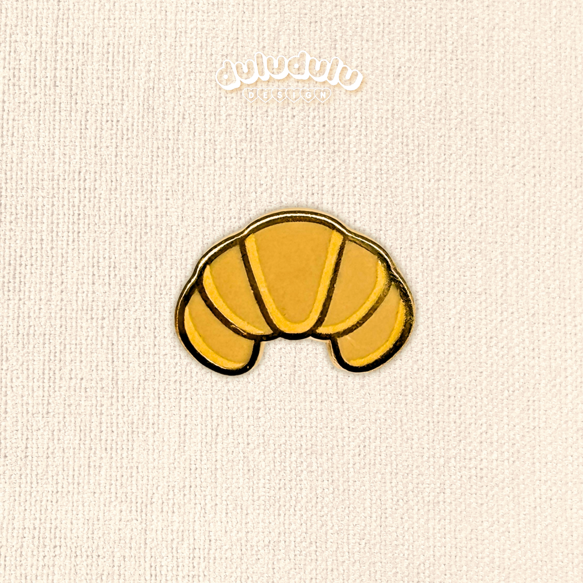 A small croissant enamel pin centered on a cream textured fabric background