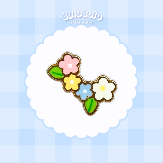 A cute enamel pin featuring four stylized flowers in pink, yellow, light blue, and white with green leaves, displayed on a white scalloped circle over a light blue checkered background.