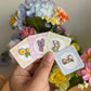 hand holding set of 4 flower mini enamel pins including sunflower, lavender, lily of the valley and flowers.