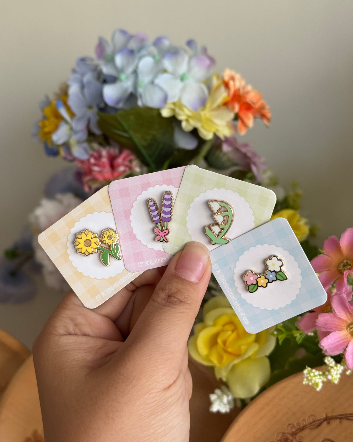 hand holding set of 4 flower mini enamel pins including sunflower, lavender, lily of the valley and flowers.
