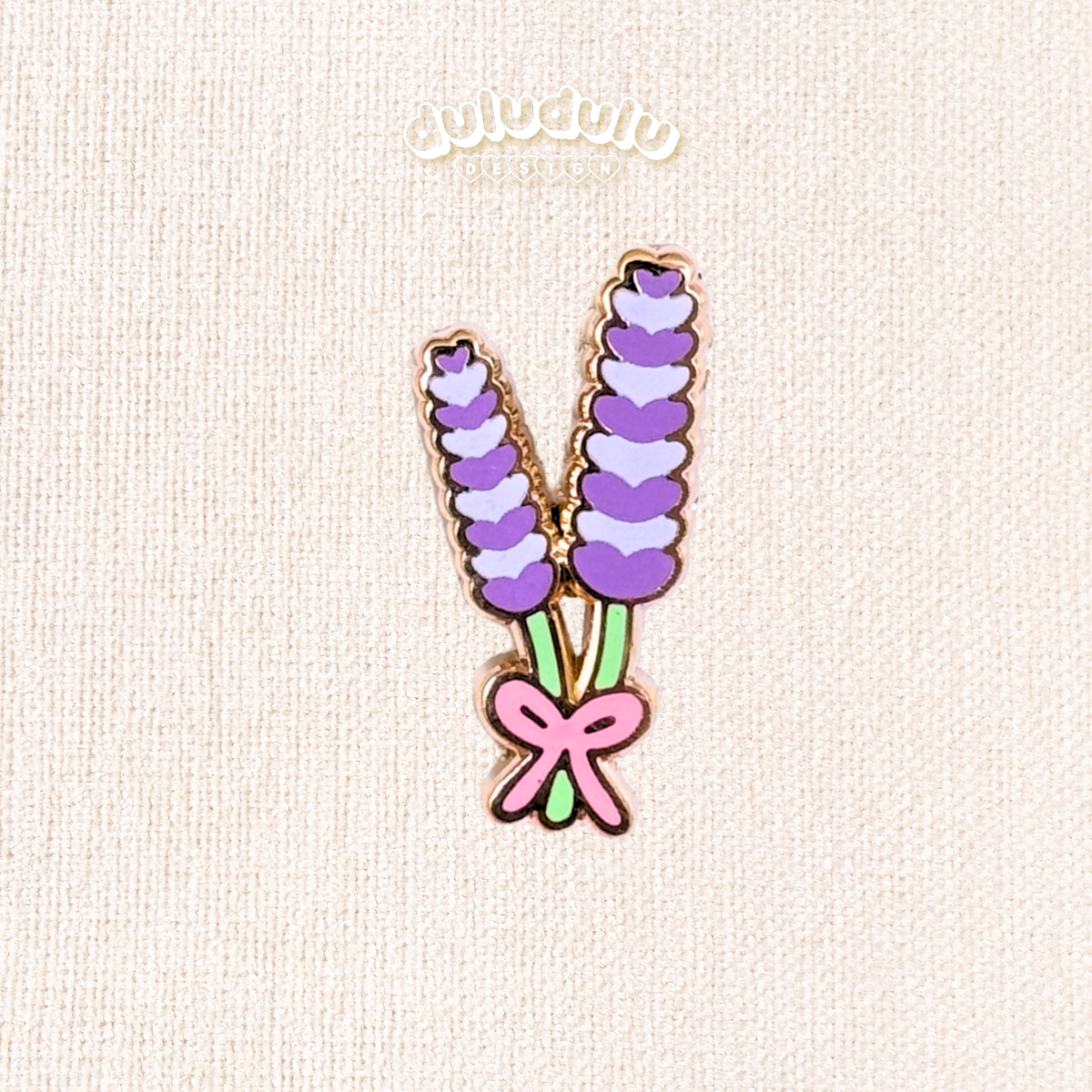 A lavender flower enamel pin displayed on a textured cream fabric background.