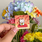 Hand holding the lucky cat mini pin under natural light, with flowers in the background