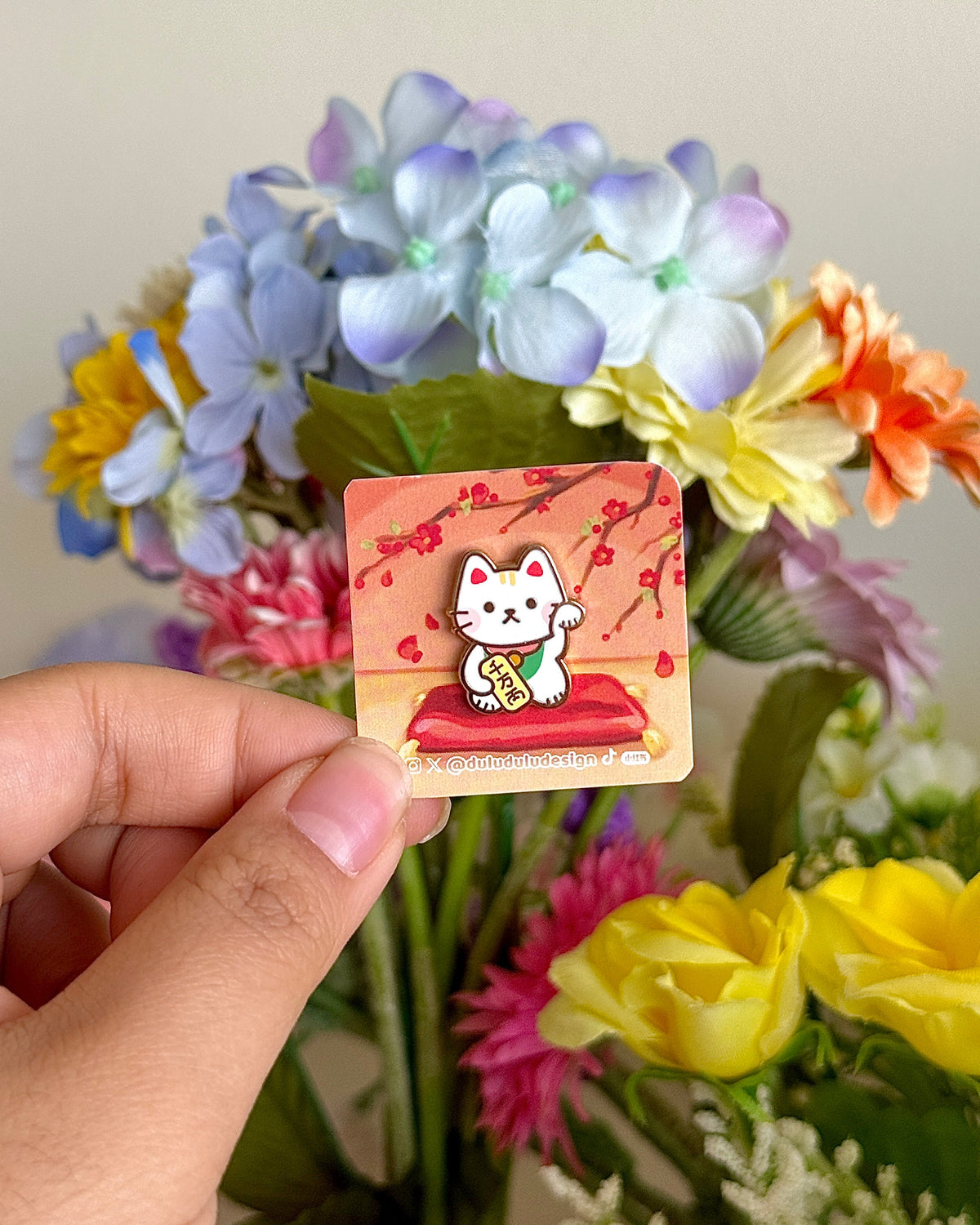 Hand holding the lucky cat mini pin under natural light, with flowers in the background