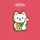 A cute white maneki-neko enamel pin with a raised paw holding a gold fortune tag, set against a textured pink background with the brand name at the top.