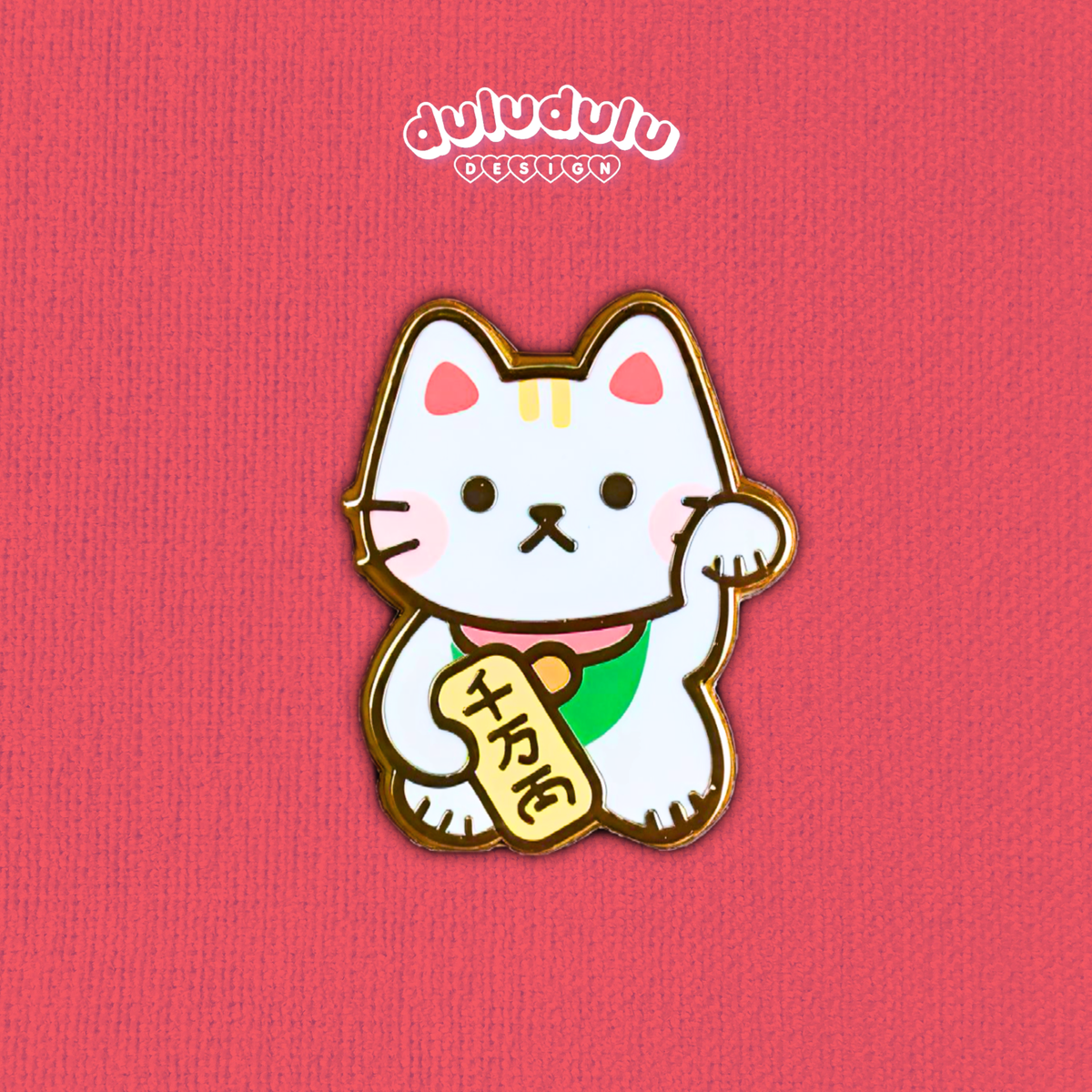 A cute white maneki-neko enamel pin with a raised paw holding a gold fortune tag, set against a textured pink background with the brand name at the top.