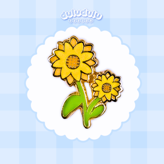 A sunflower enamel pin centered on a white scalloped circle over a light blue checkered background. 