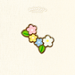 A small enamel pin shaped like a cluster of four flowers with green leaves, shown on a light cream textured background. 